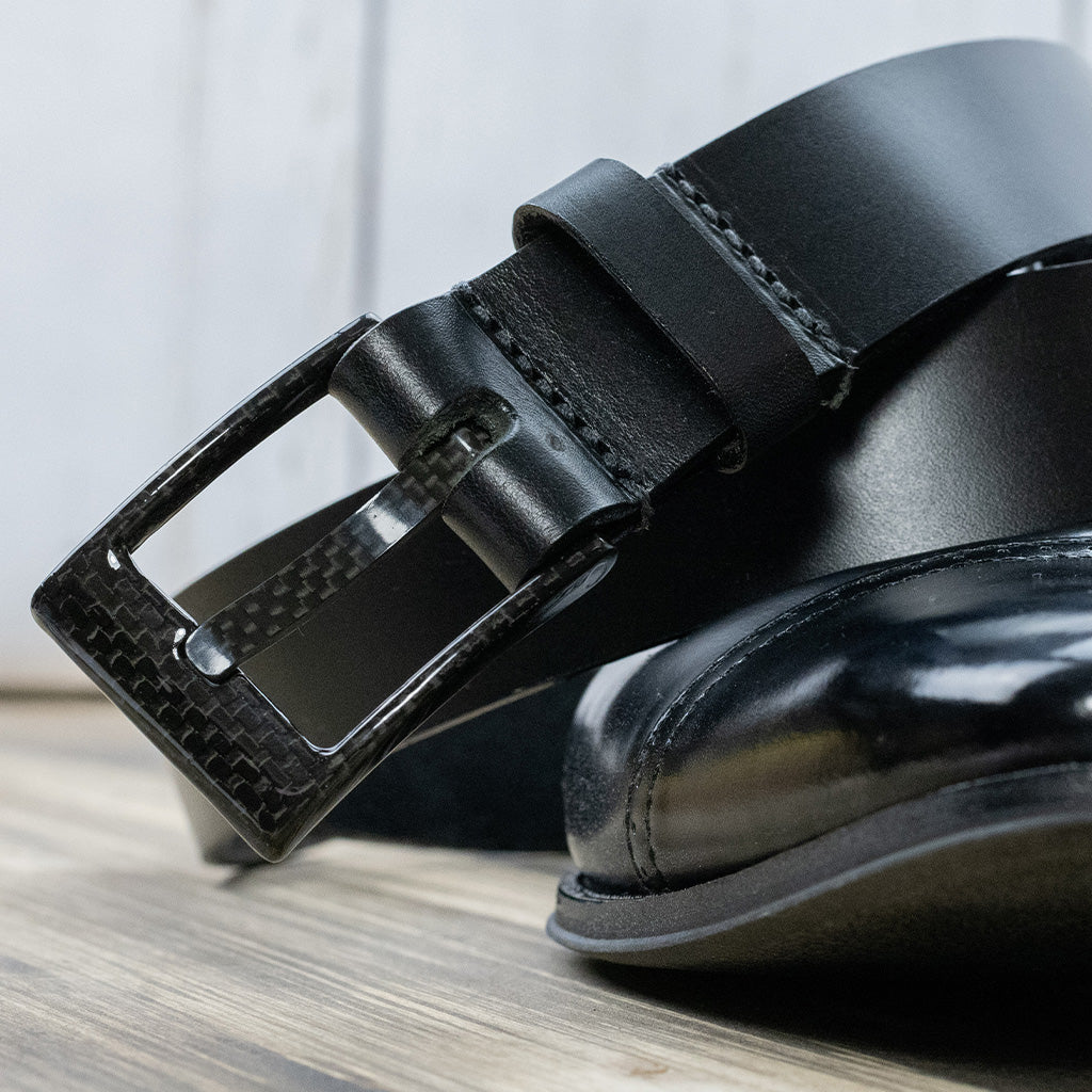 Image of The Classified Black Leather Dress Belt on top of a black dress shoe. The Classified black belt has a shiny, black leather strap with a square, black carbon fiber buckle. 