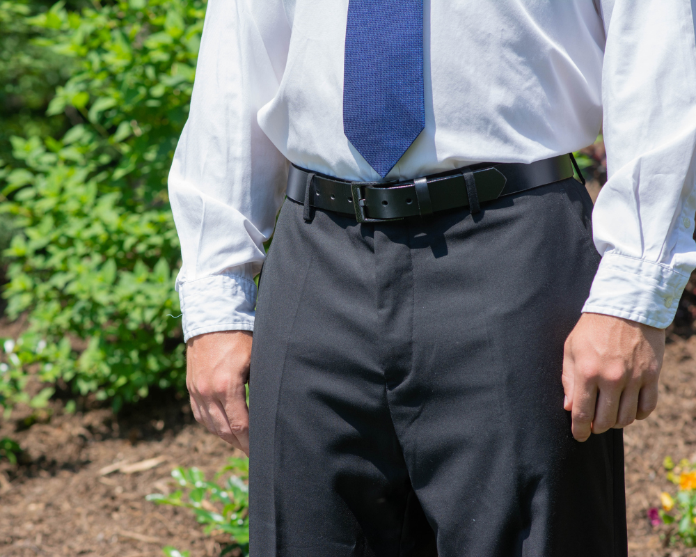 Image of a man in business attire - black slacks, blue tie, white button-up shirt with The Classified Black Leather Dress Belt. The Classified Black Belt has a solid, shiny black leather strap with a rectangular, black carbon fiber buckle.  