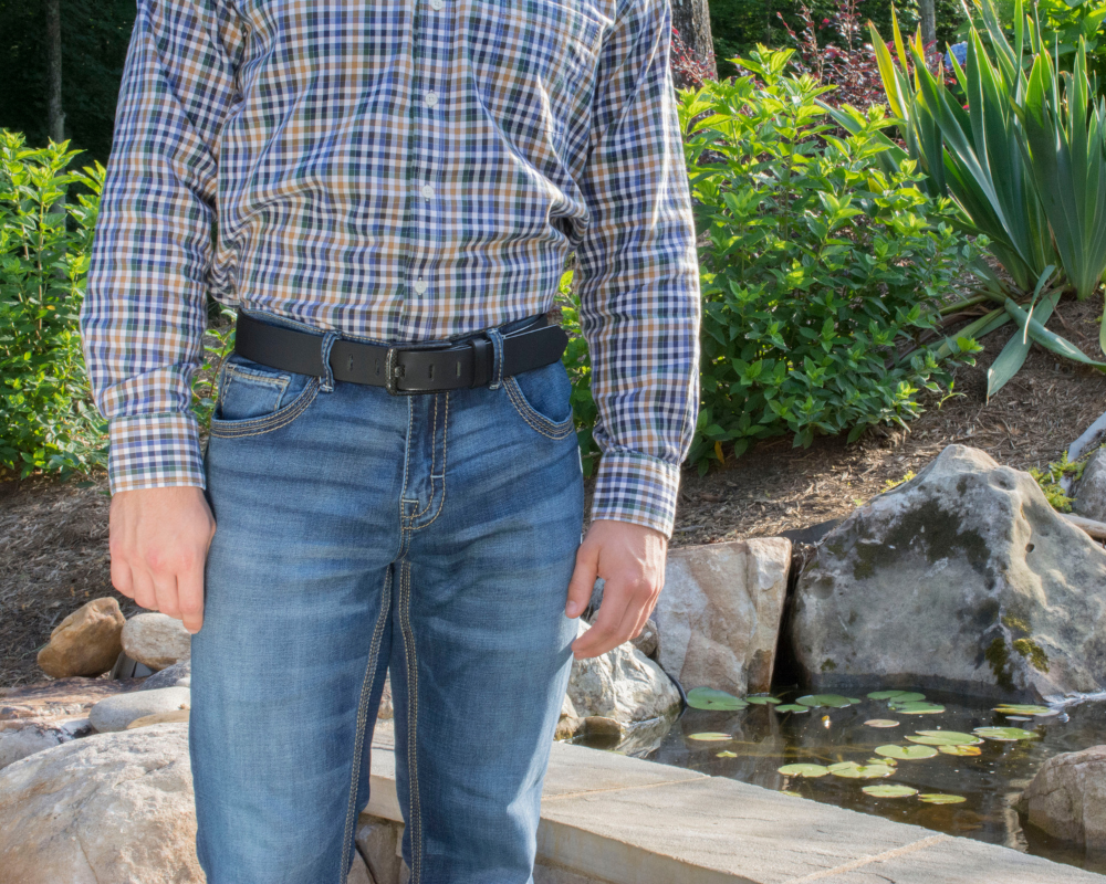 Image of a man in a nature background wearing jeans, a plaid button-up shirt and The Carbon Fiber Wide Pin Black Leather Dress Belt. The Carbon Fiber Wide Pin Belt has a shiny solid black strap with a black carbon fiber buckle that has a wide pin.