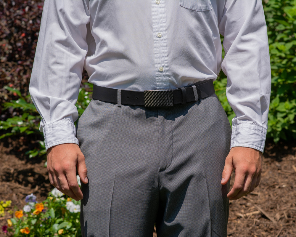 Image of a man wearing grey slacks, light white button down shirt and the CF 2.0 Black Leather Dress Belt. The CF 2.0 Black Leather Dress Belt has a solid, shiny black leather strap and a black hook buckle made of carbon fiber.