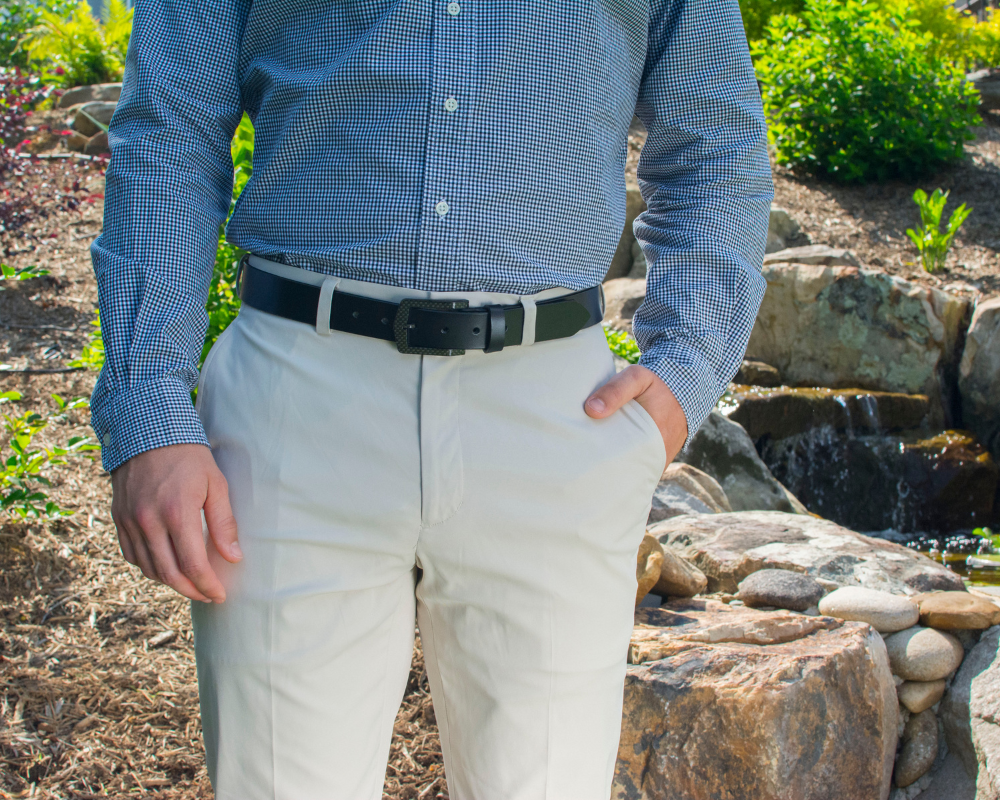 Image of a man in a natural background wearing khakis, a white/blue patterned button-up and the Stealth Black Leather Dress Belt. The Stealth Black Belt has a shiny all-black leather strap with a black carbon fiber buckle.